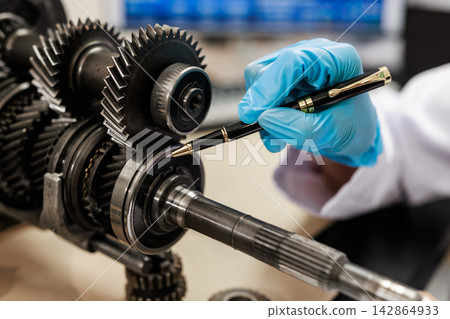 Close-up of a technician in blue gloves inspecting metal gears and a transmission shaft with a pen in an automotive repair workshop. Close-up of a technician in blue gloves inspecting metal gears and a transmission shaft with a pen in an automotive repair workshop. 142864933