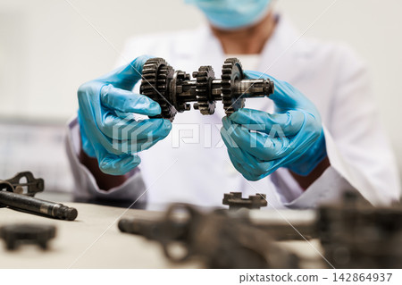 Mechanic in a white coat, face mask and blue gloves inspecting a metal gearbox shaft with gears in a workshop. Mechanic in a white coat, face mask and blue gloves inspecting a metal gearbox shaft with gears in a workshop. 142864937