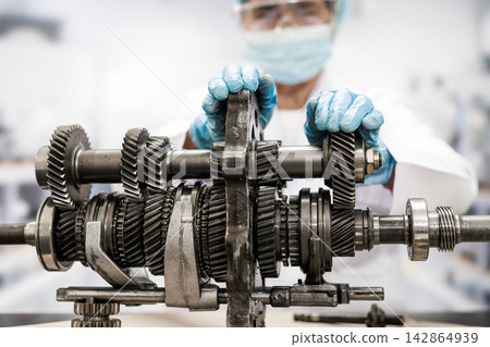 Technician wearing gloves and a face mask inspects a disassembled vehicle gearbox with metal gears and shafts in a bright repair workshop. Technician wearing gloves and a face mask inspects a disassembled vehicle gearbox with metal gears and shafts in a bright repair workshop. 142864939