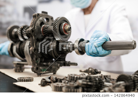 Mechanic in protective gloves repairs a vehicle gearbox, holding a metal shaft beside exposed gears and engine parts in a clean workshop. Mechanic in protective gloves repairs a vehicle gearbox, holding a metal shaft beside exposed gears and engine parts in a clean workshop. 142864940