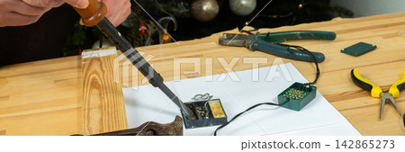 A man works on fixing a broken Christmas tree garland by soldering wires together. He uses tools and parts on a wooden table, focusing on the repair process during the holiday season, banner A man works on fixing a broken Christmas tree garland by soldering wires together. He uses tools and parts on a wooden table, focusing on the repair process during the holiday season, banner 142865273