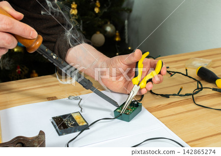 A man works on fixing a broken Christmas tree garland by soldering wires together. He uses tools and parts on a wooden table, focusing on the repair process during the holiday season A man works on fixing a broken Christmas tree garland by soldering wires together. He uses tools and parts on a wooden table, focusing on the repair process during the holiday season 142865274