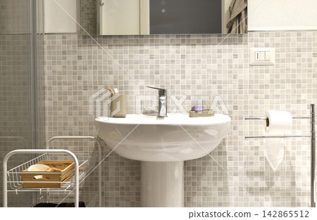 White ceramic pedestal sink with chrome faucet standing near mosaic tile wall. White ceramic pedestal sink with chrome faucet standing near mosaic tile wall. 142865512