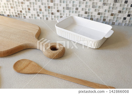 White ceramic baking dish with wooden cutting board and spoon on kitchen counter. White ceramic baking dish with wooden cutting board and spoon on kitchen counter. 142866008