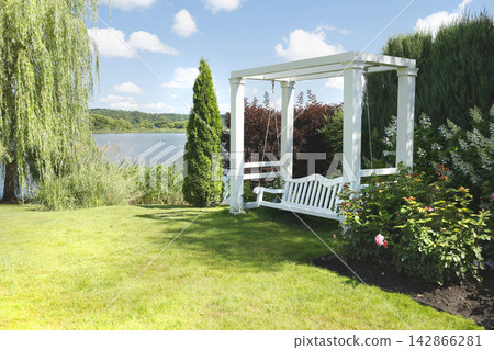 Elegant white wooden swing bench under pergola on green lakeside lawn. Luxury backyard relaxation zone featuring scenic waterfront view and beautiful landscape design. Contemporary garden decoration. Elegant white wooden swing bench under pergola on green lakeside lawn. Luxury backyard relaxation zone featuring scenic waterfront view and beautiful landscape design. Contemporary garden decoration. 142866281