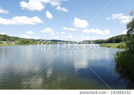 Beautiful scenic lake with calm blue water surface under fluffy clouds sky. Peaceful natural landscape featuring green forest shores and distant village houses. Tranquil countryside environment. Beautiful scenic lake with calm blue water surface under fluffy clouds sky. Peaceful natural landscape featuring green forest shores and distant village houses. Tranquil countryside environment. 142866282