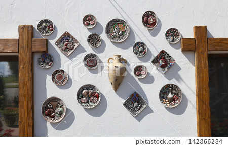 Traditional decorative ceramic plates and clay amphora hanging on white wall. Beautiful handmade pottery souvenirs featuring ethnic patterns and pomegranate illustrations. Authentic cultural interior. 142866284