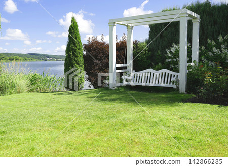 Elegant white wooden swing bench under pergola on green lakeside lawn. Luxury backyard relaxation zone featuring scenic waterfront view and beautiful landscape design. Contemporary garden decoration. 142866285