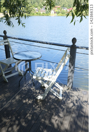 Cozy lakeside restaurant terrace with glass coffee table and white wooden chairs. Scenic waterfront wooden deck overlooking calm blue water pond and countryside village. Beautiful outdoor relaxation. Cozy lakeside restaurant terrace with glass coffee table and white wooden chairs. Scenic waterfront wooden deck overlooking calm blue water pond and countryside village. Beautiful outdoor relaxation. 142866313