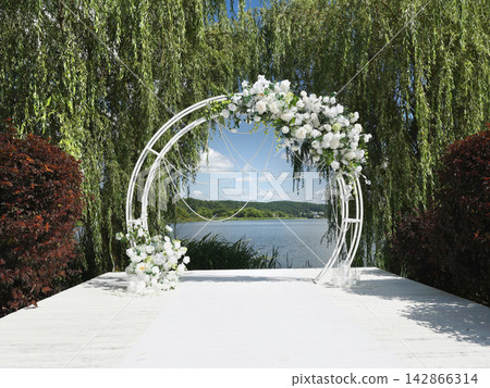 Luxury wedding venue featuring romantic floral arch on white wooden pier next to scenic lake. Elegant marriage ceremony location with exquisite white flower arrangements and crystal decorations. Luxury wedding venue featuring romantic floral arch on white wooden pier next to scenic lake. Elegant marriage ceremony location with exquisite white flower arrangements and crystal decorations. 142866314