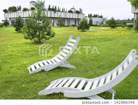 White wooden sun loungers on lush eco hotel lawn. Creative sustainable resort architecture landscaping design. Peaceful summer park garden hospitality relaxation nature background. White wooden sun loungers on lush eco hotel lawn. Creative sustainable resort architecture landscaping design. Peaceful summer park garden hospitality relaxation nature background. 142866348
