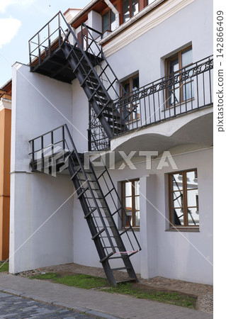 Industrial metal fire escape stairs building facade. External black iron emergency staircase structure on old renovated house wall architectural background. Industrial metal fire escape stairs building facade. External black iron emergency staircase structure on old renovated house wall architectural background. 142866409