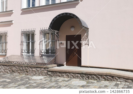 Aesthetic pastel pink building exterior cottagecore mood. Trendy minimalist facade with forged iron entrance awning and vintage windows security bars casting graphic summer shadows. Aesthetic pastel pink building exterior cottagecore mood. Trendy minimalist facade with forged iron entrance awning and vintage windows security bars casting graphic summer shadows. 142866735