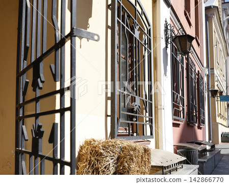 Forged iron window bars gate foreground photo. Vintage metal security grills and antique street lantern on colorful building facade wall. European old town street perspective side view daylight. Forged iron window bars gate foreground photo. Vintage metal security grills and antique street lantern on colorful building facade wall. European old town street perspective side view daylight. 142866770