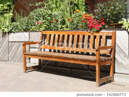 Wooden park bench garden photo. Empty varnished timber seat stands on brick pavement in front of blooming flowerbed with red orange blossoms. Classical outdoor backyard patio furniture design closeup Wooden park bench garden photo. Empty varnished timber seat stands on brick pavement in front of blooming flowerbed with red orange blossoms. Classical outdoor backyard patio furniture design closeup 142866771