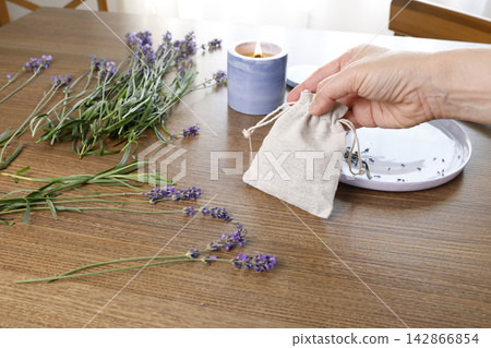 Woman hand holding small linen sachet filled with dried lavender. Scented candle burning on wooden table near herbal bouquet arrangement. Handmade aromatherapy pouch craft workshop relaxation lifestyl Woman hand holding small linen sachet filled with dried lavender. Scented candle burning on wooden table near herbal bouquet arrangement. Handmade aromatherapy pouch craft workshop relaxation lifestyl 142866854