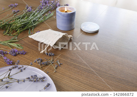 Linen sachet bag purple plate dried lavender buds burning candle wooden table backdrop. Side view craft hobby handmade herbal pillow process copy space. Creative organic artisan workshop concept. Linen sachet bag purple plate dried lavender buds burning candle wooden table backdrop. Side view craft hobby handmade herbal pillow process copy space. Creative organic artisan workshop concept. 142866872