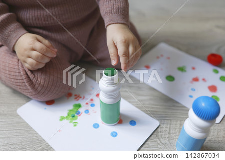 Toddler hands close-up touching green dot marker tip on drawing paper. Creative educational activity for fine motor skills development at home. Real indoor lifestyle cropped macro view Toddler hands close-up touching green dot marker tip on drawing paper. Creative educational activity for fine motor skills development at home. Real indoor lifestyle cropped macro view 142867034
