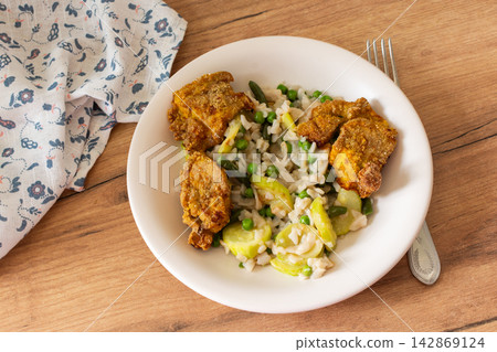 A bowl contains gluten-free pasta with baked zucchini, peas, and green beans alongside deep-fried chicken nuggets. This meal is served on a wooden table with bread in the background A bowl contains gluten-free pasta with baked zucchini, peas, and green beans alongside deep-fried chicken nuggets. This meal is served on a wooden table with bread in the background 142869124