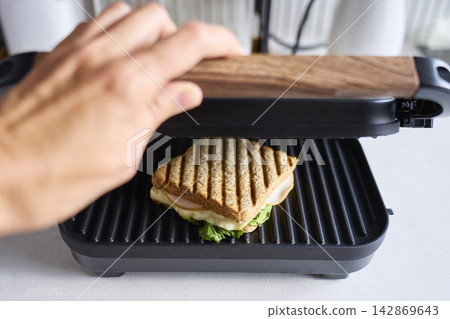 Person lowering sandwich press lid over toasted sandwich with cheese, lettuce and sliced meat Person lowering sandwich press lid over toasted sandwich with cheese, lettuce and sliced meat 142869643
