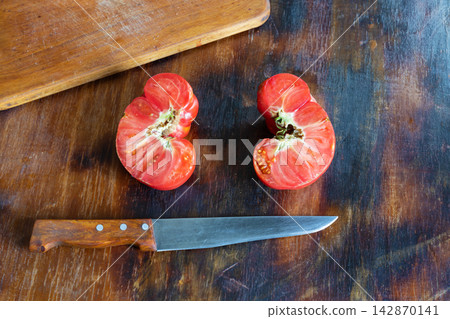 Two halves of unusually shaped, ugly red tomato lie on rustic wooden table after being cut open with kitchen knife. Cut surfaces reveal fleshy interior, seed chambers, and irregular natural structure Two halves of unusually shaped, ugly red tomato lie on rustic wooden table after being cut open with kitchen knife. Cut surfaces reveal fleshy interior, seed chambers, and irregular natural structure 142870141