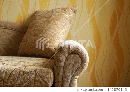 Close-up view of cozy upholstered sofa with decorative cushions and rounded armrest in warm brown and golden tones. Textured fabric, geometric patterns, soft lighting create comfortable room interior Close-up view of cozy upholstered sofa with decorative cushions and rounded armrest in warm brown and golden tones. Textured fabric, geometric patterns, soft lighting create comfortable room interior 142870143