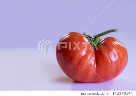 Naturally misshapen ripe red tomato with asymmetrical form and pronounced lobes, on soft lavender background. Natural variation and imperfection in agricultural produce, with copy space for design Naturally misshapen ripe red tomato with asymmetrical form and pronounced lobes, on soft lavender background. Natural variation and imperfection in agricultural produce, with copy space for design 142870144