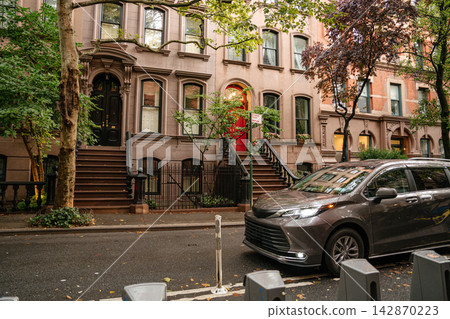 Wide view of classic New York brownstone townhouses along a tree lined residential street with parked car in the foreground. Historic stone facades and stoop staircases define the iconic urban Wide view of classic New York brownstone townhouses along a tree lined residential street with parked car in the foreground. Historic stone facades and stoop staircases define the iconic urban 142870223