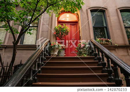 Elegant red door entrance of a historic New York City brownstone with classic stoop stairs and black iron railings. Flower pots and greenery decorate the facade, creating a charming residential urban Elegant red door entrance of a historic New York City brownstone with classic stoop stairs and black iron railings. Flower pots and greenery decorate the facade, creating a charming residential urban 142870233