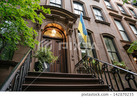 Low angle perspective of a historic New York brownstone townhouse showing detailed stone facade, wooden door, and iron staircase. A Ukrainian flag is displayed near the entrance, adding cultural and Low angle perspective of a historic New York brownstone townhouse showing detailed stone facade, wooden door, and iron staircase. A Ukrainian flag is displayed near the entrance, adding cultural and 142870235
