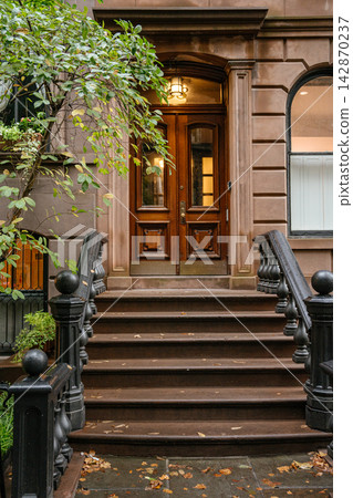 A detailed view of a classic New York brownstone entrance with carved wooden double doors and black iron railings along the steps. Green tree branches frame the historic residential facade in an urban A detailed view of a classic New York brownstone entrance with carved wooden double doors and black iron railings along the steps. Green tree branches frame the historic residential facade in an urban 142870237