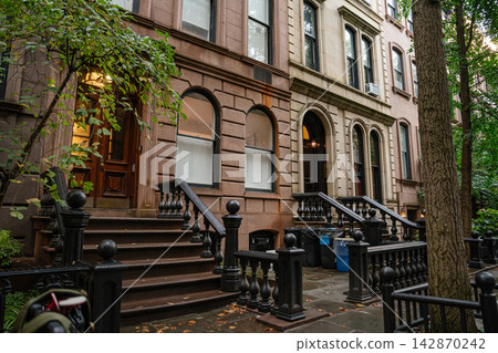 A row of classic brownstone townhouses with stone staircases and iron railings on a quiet New York City street. The residential buildings are framed by trees and urban greenery in a peaceful 142870242