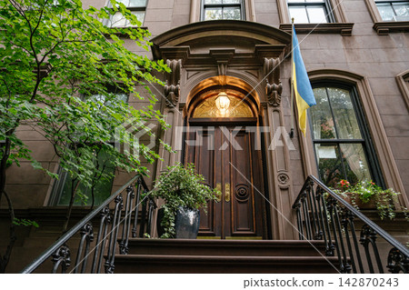 A classic New York brownstone townhouse entrance with ornate wooden double doors, stone arch details, and a Ukrainian flag hanging beside the doorway. The historic city building is surrounded by green A classic New York brownstone townhouse entrance with ornate wooden double doors, stone arch details, and a Ukrainian flag hanging beside the doorway. The historic city building is surrounded by green 142870243