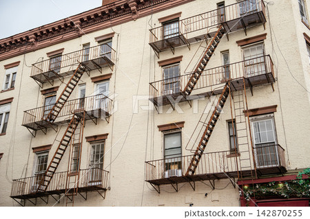 A historic white brick apartment building with metal fire escapes on the facade. The urban architecture shows classic New York style residential design. A historic white brick apartment building with metal fire escapes on the facade. The urban architecture shows classic New York style residential design. 142870255