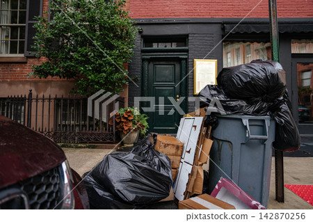 Black garbage bags and cardboard boxes stacked beside a trash bin on a New York City sidewalk. Urban street scene shows residential building entrance and everyday city life. Black garbage bags and cardboard boxes stacked beside a trash bin on a New York City sidewalk. Urban street scene shows residential building entrance and everyday city life. 142870256
