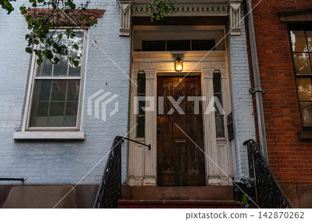 A weathered wooden entrance door set in a classic New York brownstone facade with decorative white trim. Warm porch light and brick exterior create a cozy residential urban atmosphere. A weathered wooden entrance door set in a classic New York brownstone facade with decorative white trim. Warm porch light and brick exterior create a cozy residential urban atmosphere. 142870262