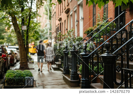 A couple walks along a tree lined New York City brownstone street with red brick buildings and black iron stair railings. Wet sidewalk, parked cars, and greenery create a cozy urban neighborhood A couple walks along a tree lined New York City brownstone street with red brick buildings and black iron stair railings. Wet sidewalk, parked cars, and greenery create a cozy urban neighborhood 142870270