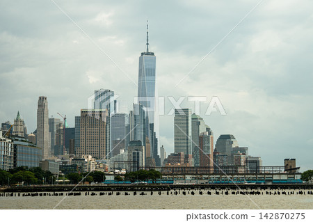 One World Trade Center rises prominently above the Lower Manhattan skyline. Modern skyscrapers line the Hudson River waterfront under a cloudy sky in New York City. One World Trade Center rises prominently above the Lower Manhattan skyline. Modern skyscrapers line the Hudson River waterfront under a cloudy sky in New York City. 142870275
