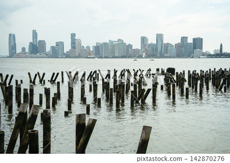 Old wooden pier pilings stand in the Hudson River with the Jersey City skyline in the distance. Modern skyscrapers rise above the waterfront under soft cloudy skies. Old wooden pier pilings stand in the Hudson River with the Jersey City skyline in the distance. Modern skyscrapers rise above the waterfront under soft cloudy skies. 142870276