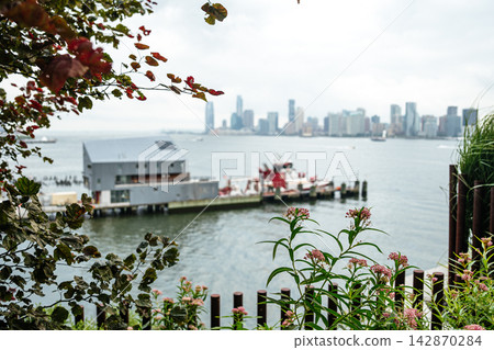 The Hudson River and Jersey City skyline are framed by green plants and pink flowers in the foreground. A waterfront pier and fireboat add urban detail to the soft, cloudy cityscape. The Hudson River and Jersey City skyline are framed by green plants and pink flowers in the foreground. A waterfront pier and fireboat add urban detail to the soft, cloudy cityscape. 142870284