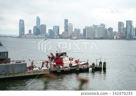 A red fireboat is docked along the Hudson River with the Jersey City skyline rising in the background. Modern skyscrapers and waterfront piers create a dramatic urban coastal scene under cloudy skies. A red fireboat is docked along the Hudson River with the Jersey City skyline rising in the background. Modern skyscrapers and waterfront piers create a dramatic urban coastal scene under cloudy skies. 142870285