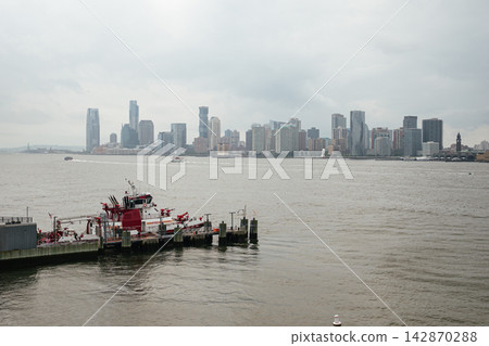 A red fireboat is docked along the Hudson River with a modern city skyline visible in the distance. Boats and waterfront infrastructure highlight urban transportation and maritime activity. A red fireboat is docked along the Hudson River with a modern city skyline visible in the distance. Boats and waterfront infrastructure highlight urban transportation and maritime activity. 142870288