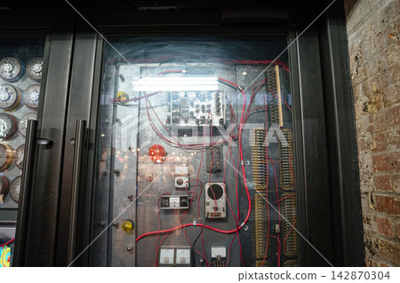 Close view of an old industrial electrical control panel with red wires, switches, and analog gauges behind glass. Metallic textures and exposed brick walls create a retro mechanical atmosphere. Close view of an old industrial electrical control panel with red wires, switches, and analog gauges behind glass. Metallic textures and exposed brick walls create a retro mechanical atmosphere. 142870304