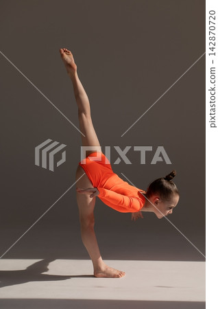 A little Caucasian girl gymnast in an orange leotard demonstrates her skills on a white background. A little Caucasian girl gymnast in an orange leotard demonstrates her skills on a white background. 142870720