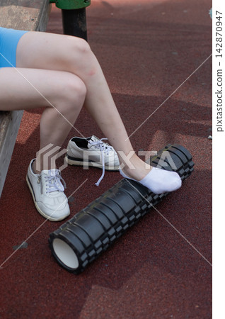 A Caucasian woman uses a foam roller to stretch her feet on a sports field. Close-up of her feet. 142870947