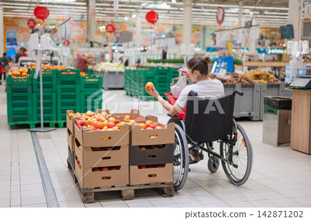 A Caucasian woman in a wheelchair buys apples at a supermarket. 142871202