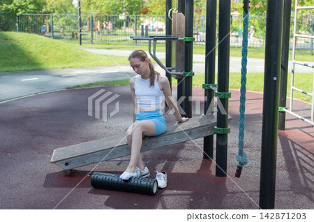 A Caucasian woman stretches her feet with a foam roller at a sports ground. A Caucasian woman stretches her feet with a foam roller at a sports ground. 142871203