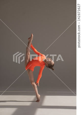 A little Caucasian girl gymnast in an orange leotard demonstrates her skills on a white background. A little Caucasian girl gymnast in an orange leotard demonstrates her skills on a white background. 142871627