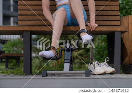 A woman uses Pedal Exercise Machine for self-rehabilitation outdoors. Close-up of feet. A woman uses Pedal Exercise Machine for self-rehabilitation outdoors. Close-up of feet. 142871682