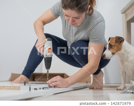 A Caucasian woman uses a screwdriver to tighten a screw while assembling a cabinet. A Caucasian woman uses a screwdriver to tighten a screw while assembling a cabinet. 142871979